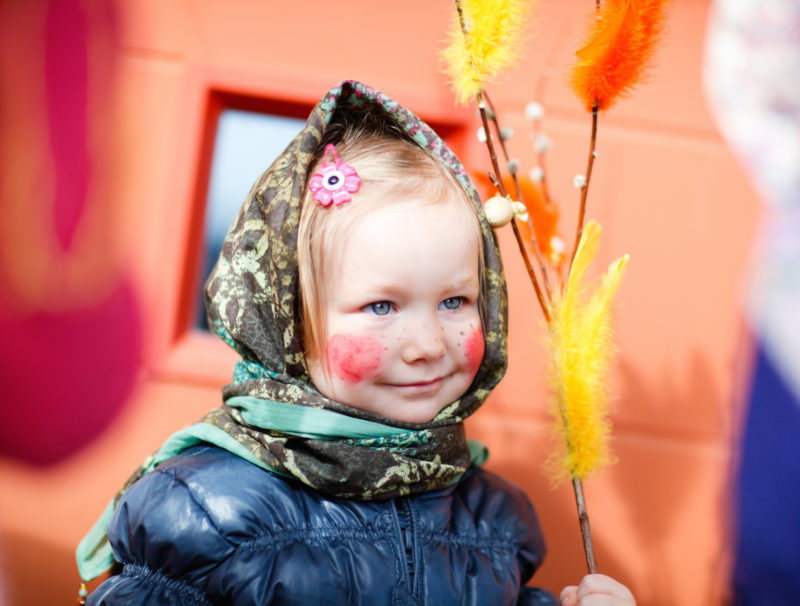 Liten påskhäxa med målade kinder, an halsduk knuten kring håret och videkvist i handen.